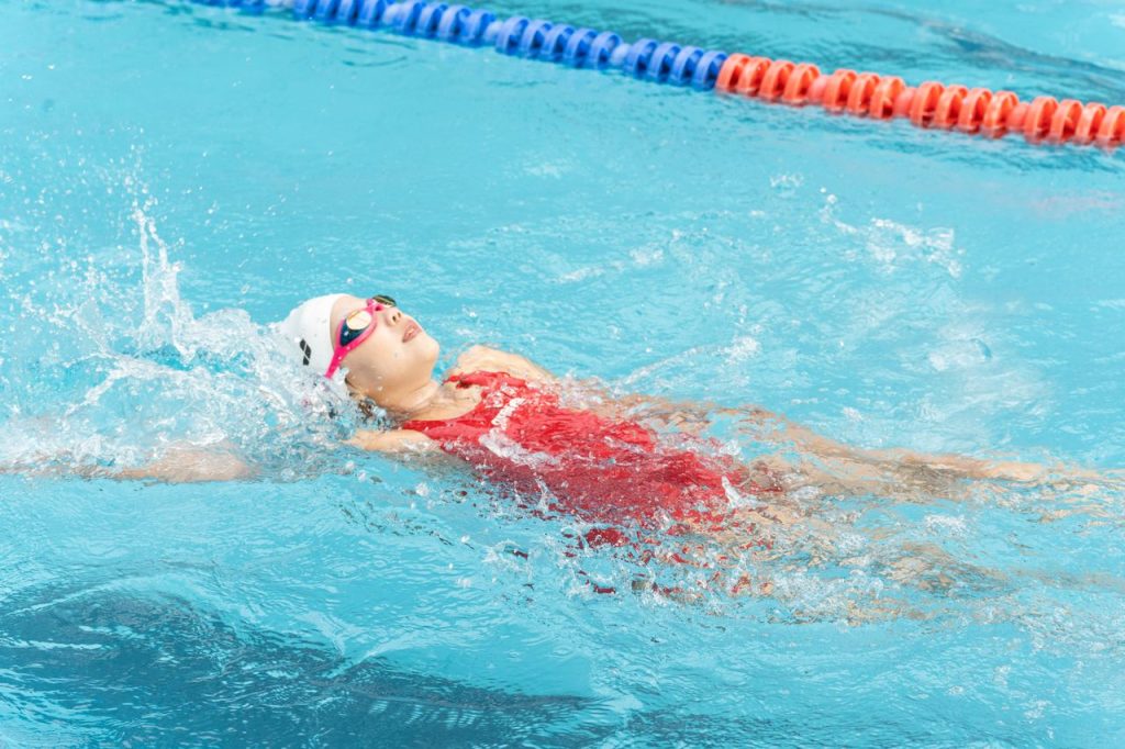 Young swimmer practicing backstroke in a swimming pool, wearing goggles and swim cap.
